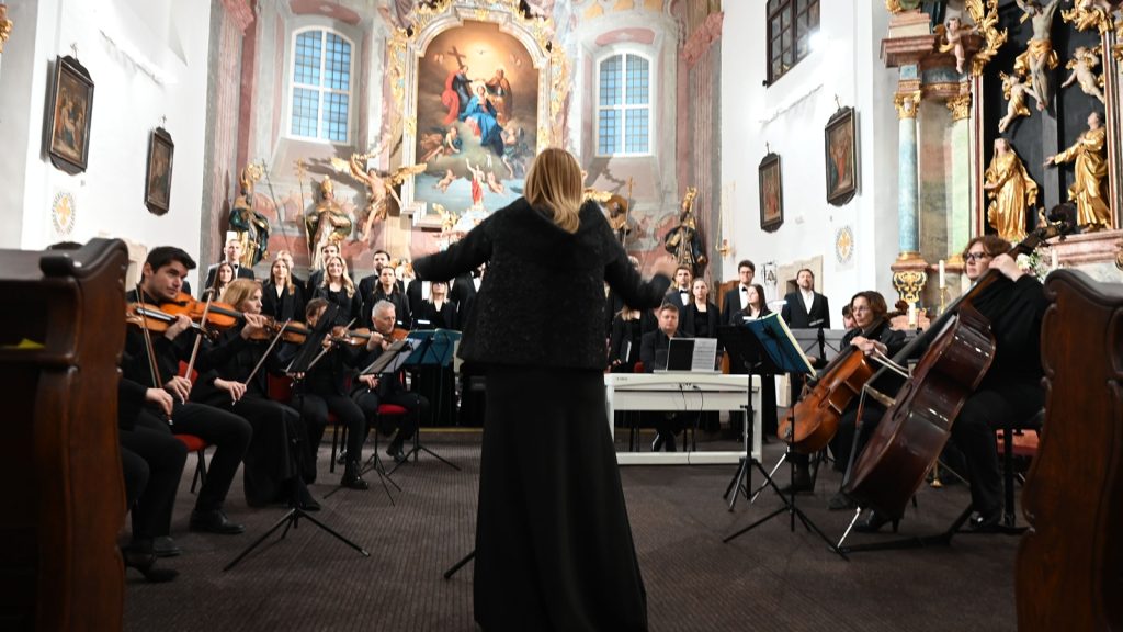 Conductor leads an orchestra and choir performing in an ornate church sanctuary.
