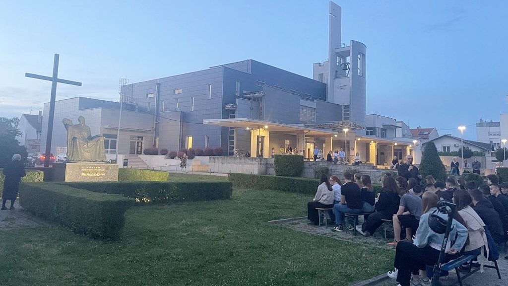 Outdoor gathering beside a modern building featuring a large cross sculpture and a seated statue on a pedestal; attendees sit on benches nearby.