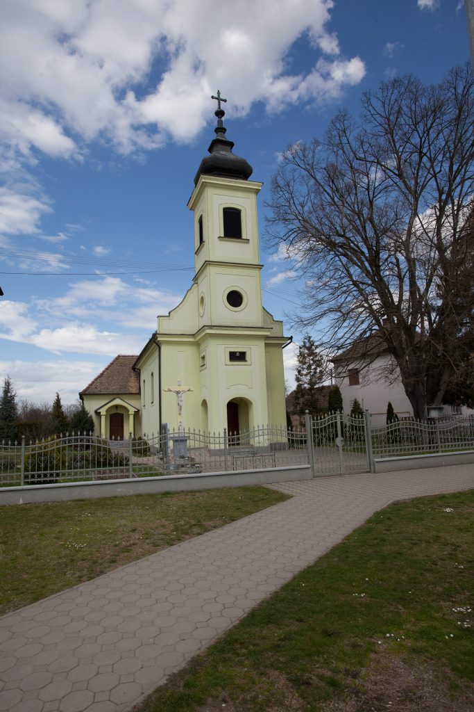 Front view of a pale yellow church with a tall steeple and cross on top, set against a blue sky with bare trees nearby.