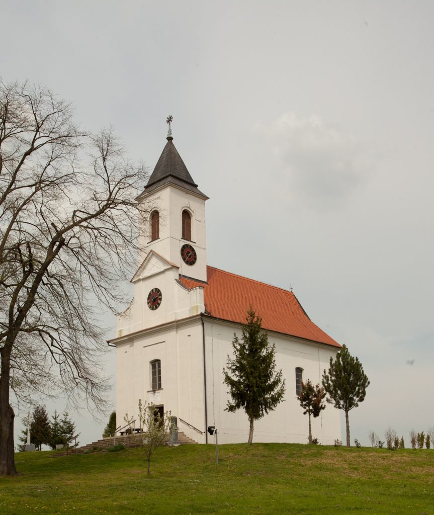 White church with a tall steeple, red-tiled roof, and clock faces, set on a grassy hill with leafless trees nearby.