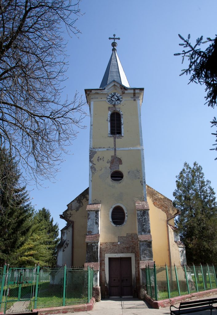Old yellow church steeple with clock, topped by a cross, set against a blue sky and surrounding trees.