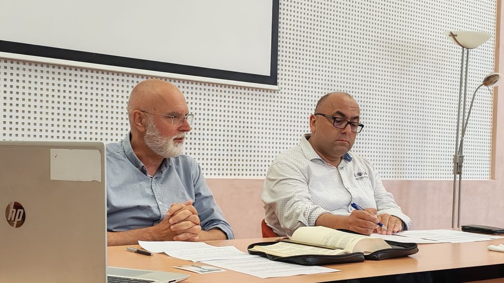 Two men sit at a conference table during a meeting; the left man has a white beard and blue shirt, the right is writing beside an open Bible on the table.