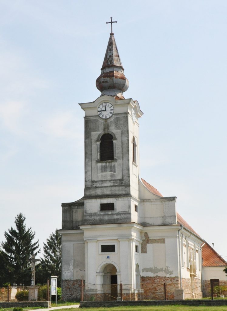 Old church with a tall clock tower and onion-shaped steeple topped by a cross against a blue sky.
