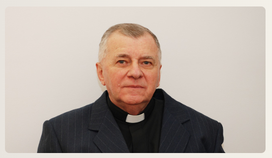 Older man wearing a clerical collar and pinstripe suit looks at the camera.
