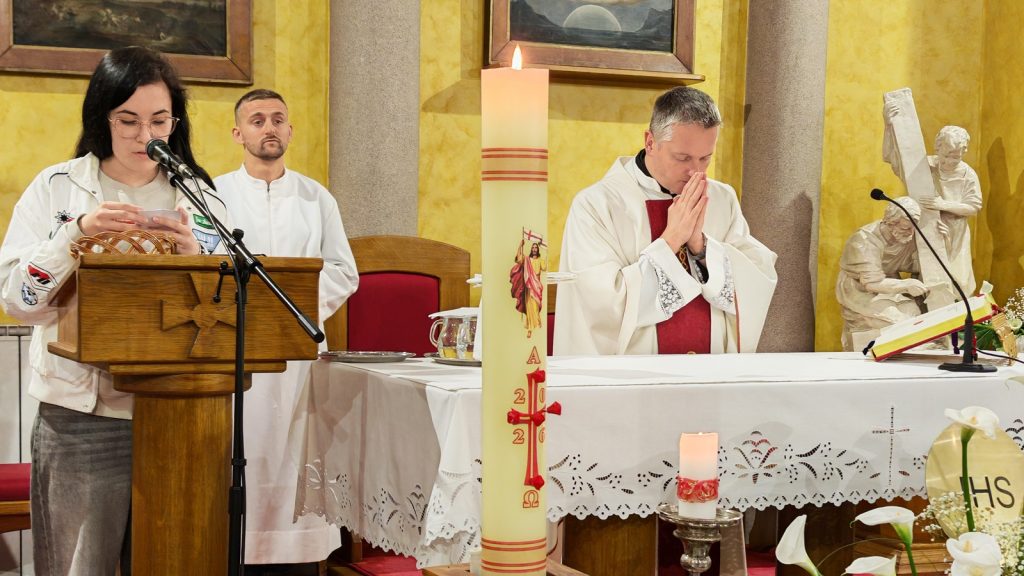 Woman at a wooden lectern reads into a microphone beside a priest praying at the altar.