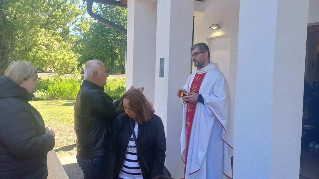 Priest in white robes blessing two women and a man outdoors near a white building with columns and greenery in the background.