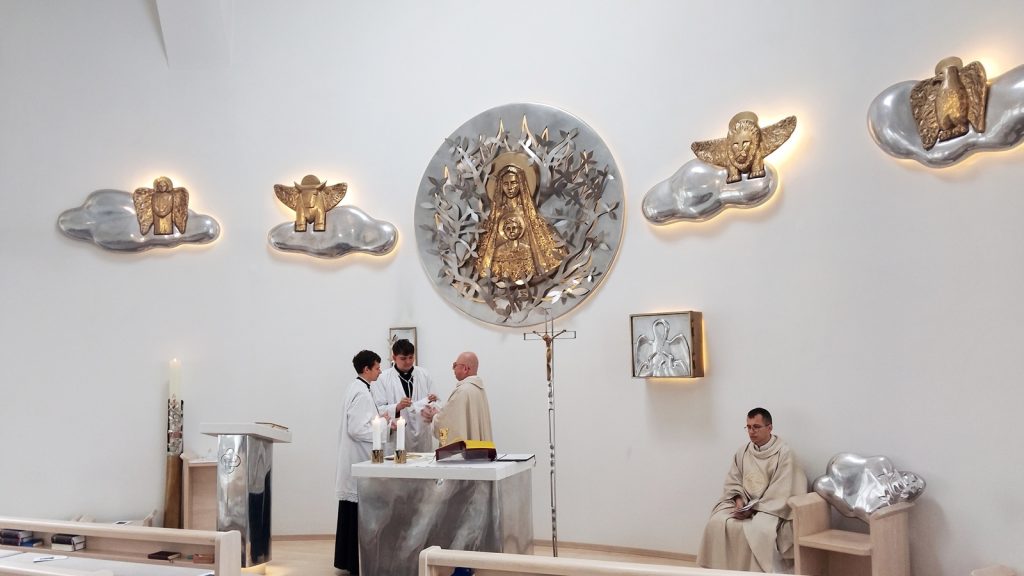Priests at an altar in a bright chapel, with a large circular Mary-and-child sculpture on the wall.