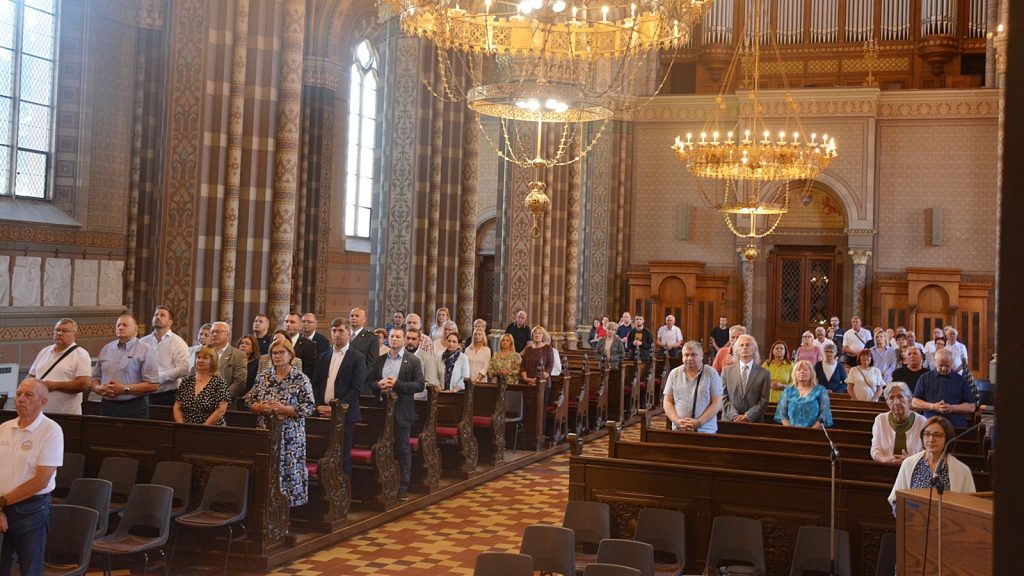Diverse group of people standing in wooden pews inside a grand, ornately decorated church with chandeliers and patterned walls.