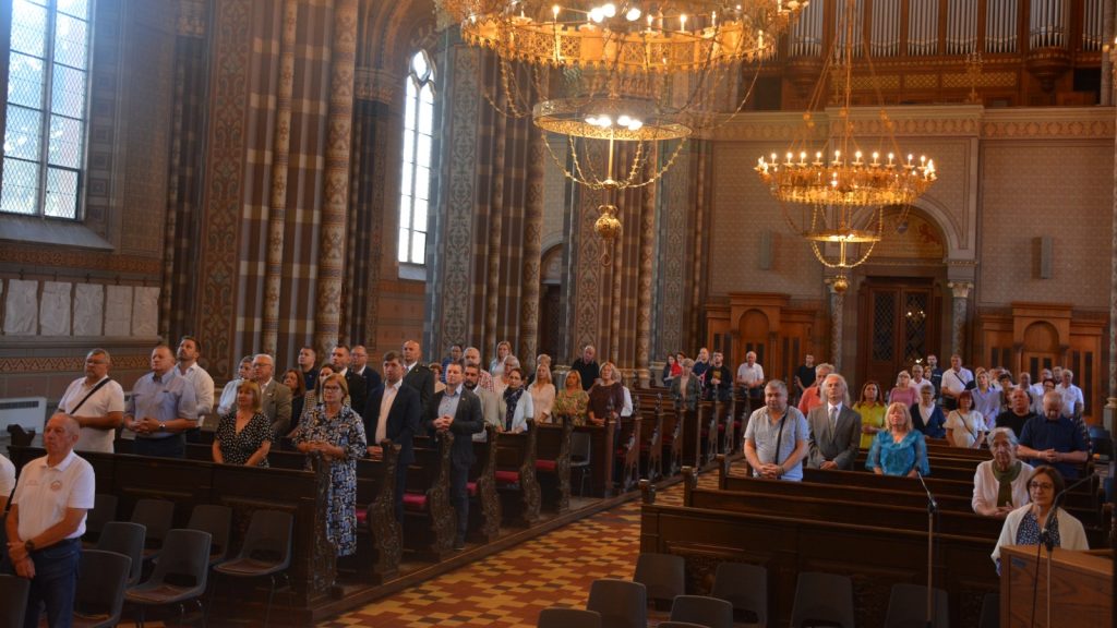 Group of people standing in pews inside a decorated cathedral with chandeliers and tall stained-glass windows.