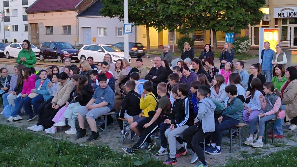Crowd of children and adults seated on benches for an outdoor community event in a town square.