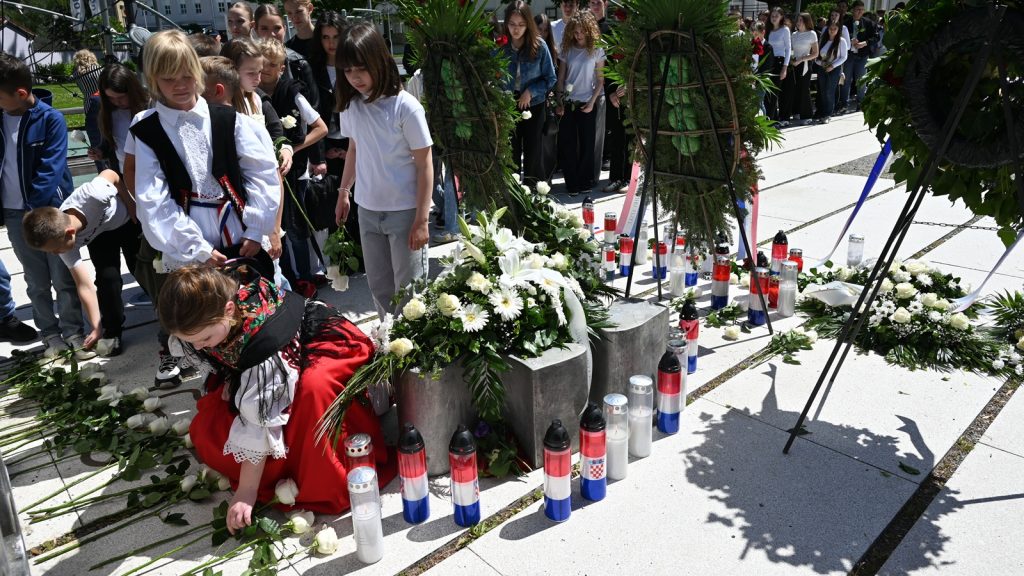 Group of children near a memorial laying white roses and candles, with wreaths and greenery in the background.