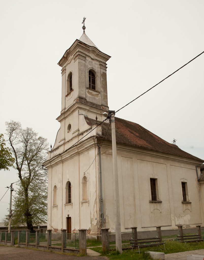 White, weathered church with a tall square bell tower topped by a small steeple, set against a pale sky; a street lamp and power line cross the foreground.