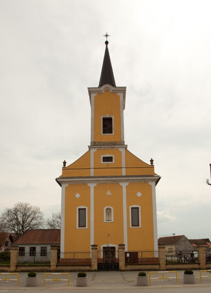 Front view of a yellow church with white trim and a tall dark steeple topped by a cross, in a town square.