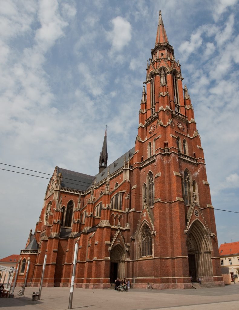 Red-brick Gothic church with a tall ornate steeple and arched windows against a blue sky.