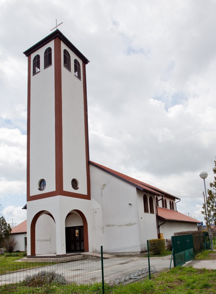 White church with a tall red-trimmed bell tower and cross, against a cloudy sky.