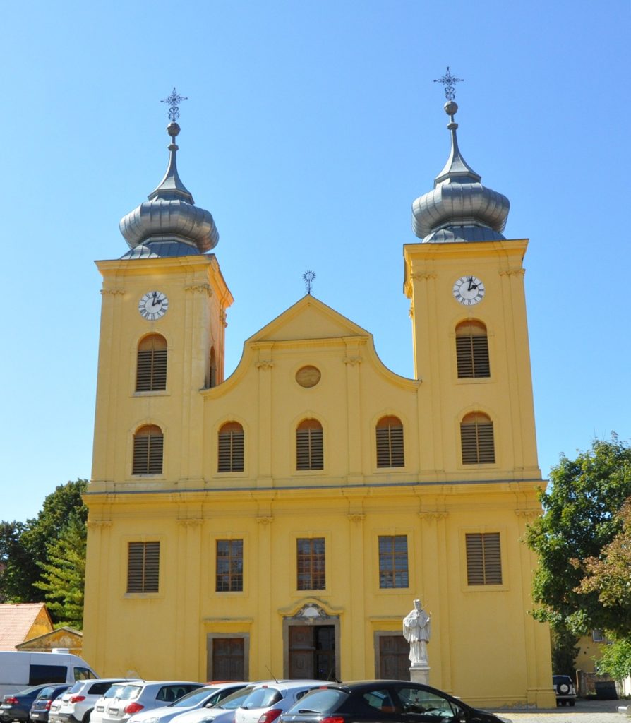 Bright yellow church with twin clock towers and decorative domes against a blue sky, with a statue and parked cars in front.