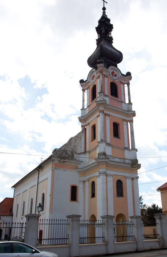 Church tower with peach and white façade, clock faces, and a black onion dome against a cloudy sky.