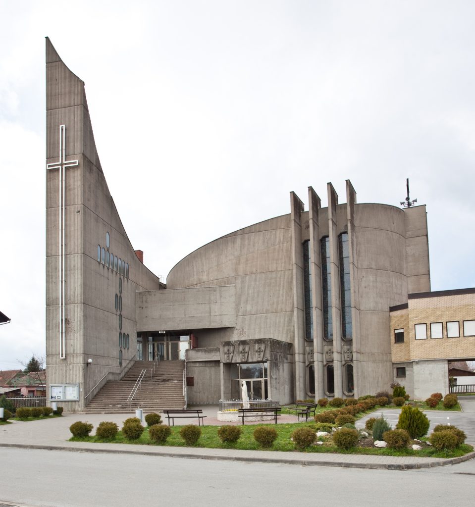 Concrete church building with a tall triangular tower and illuminated cross on the left, steps leading to the entrance, and a small courtyard with benches and shrubs in front.