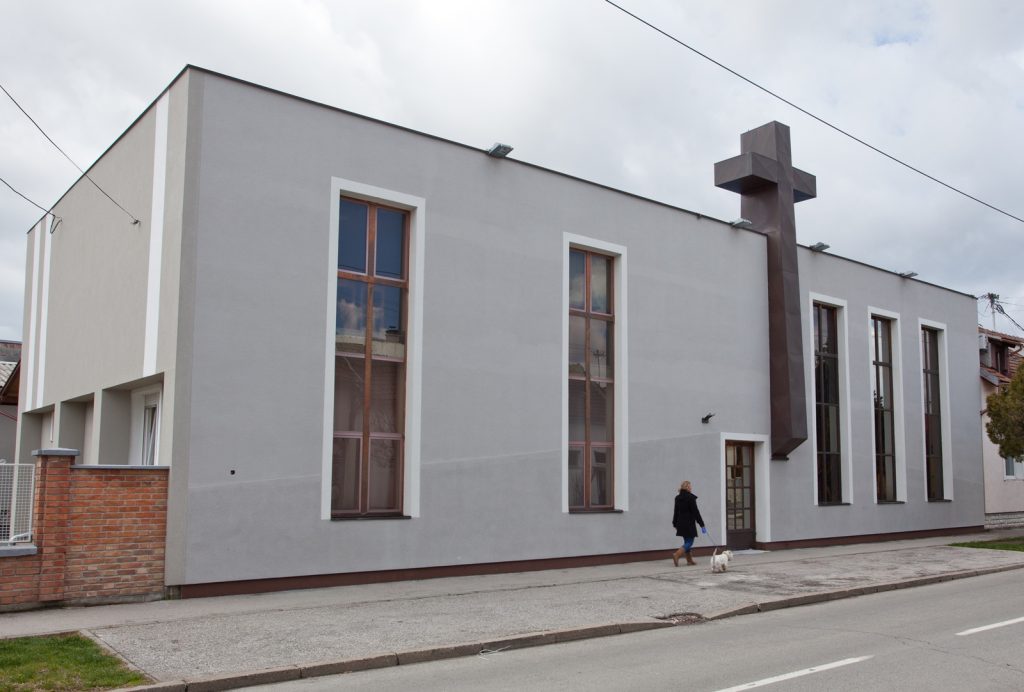 Modern gray building with tall rectangular windows and a large cross-shaped structure on the roof; a person walks a small dog on the sidewalk.