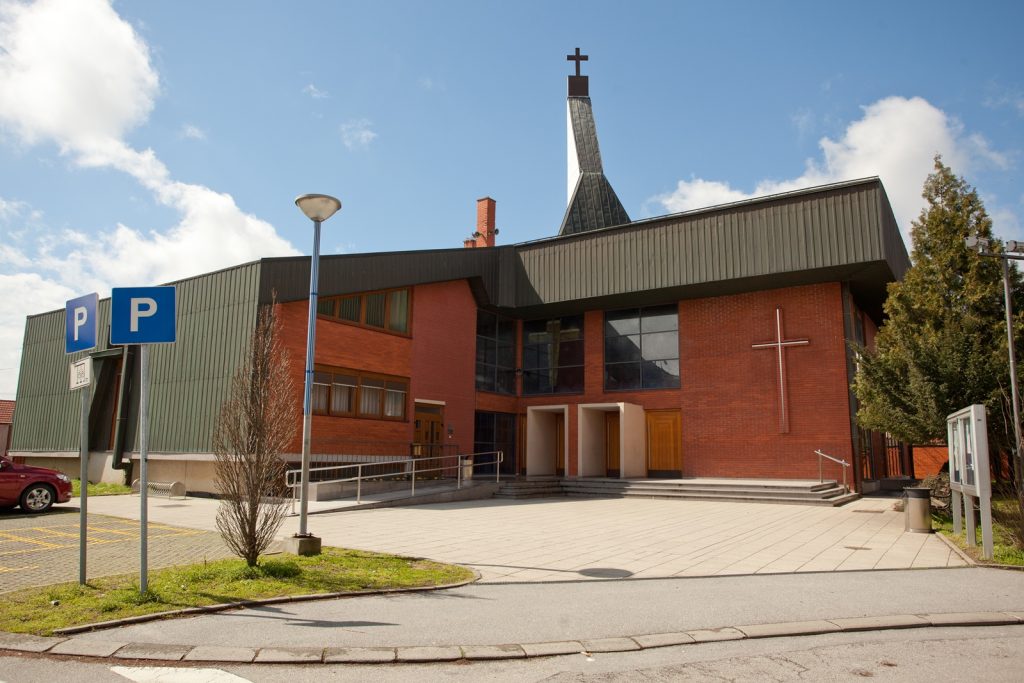 Modern brick church with a cross on the wall, large windows, and a tall steeple against a blue sky; parking signs in the foreground.