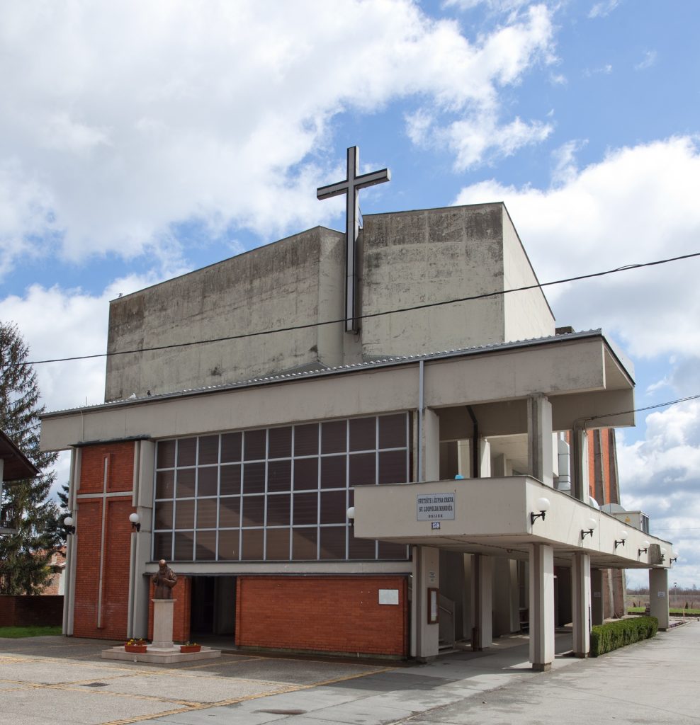 Modern church building with a metal cross atop the roof against a blue sky with clouds, brick and concrete facade nearby statue pedestal.