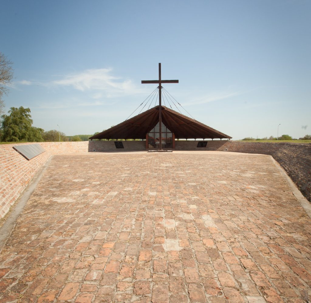 Open brick memorial with a tall cross atop a wooden pavilion in an open plaza under a clear sky.