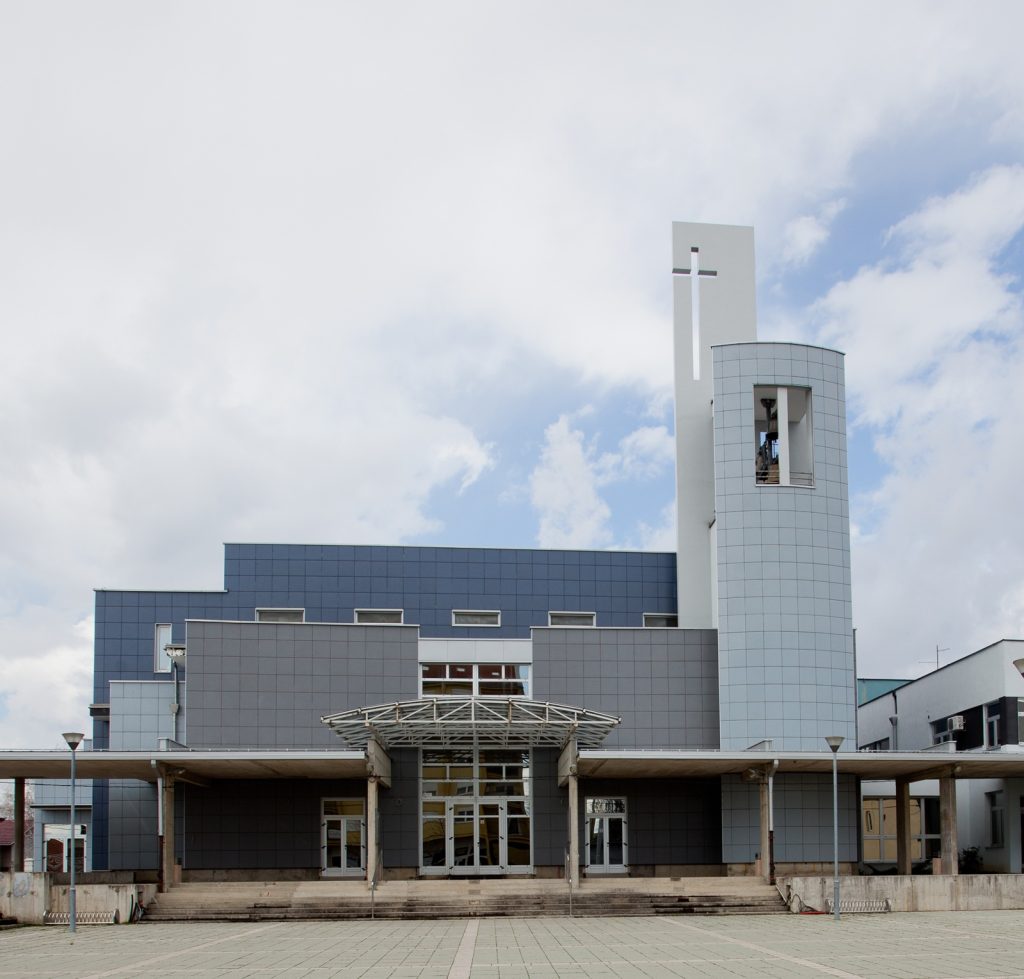 Modern gray-blue building with a tall white tower featuring a cross-shaped cutout against a cloudy sky.