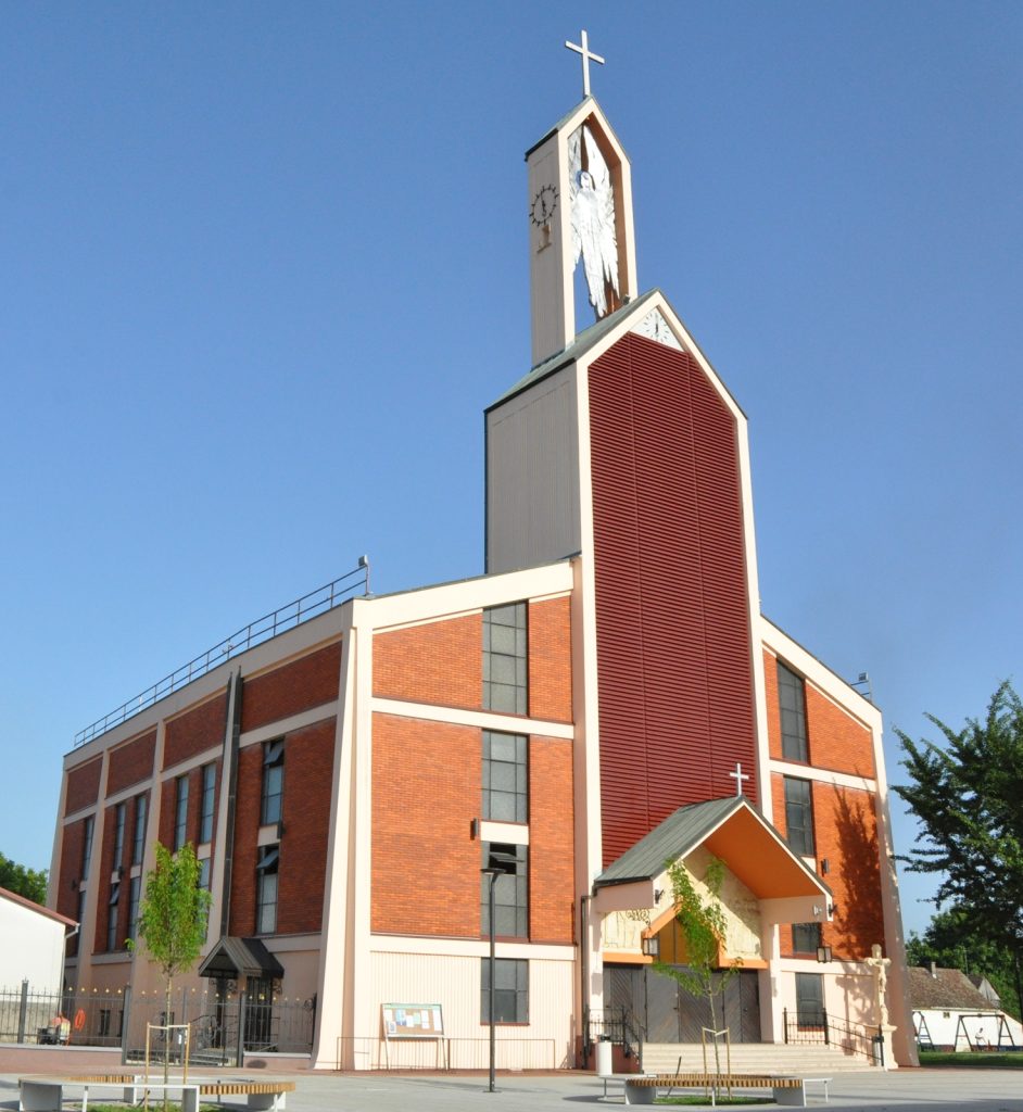 Modern brick church building with a tall steeple, cross on top, and large red louvered vent panels against a clear blue sky.