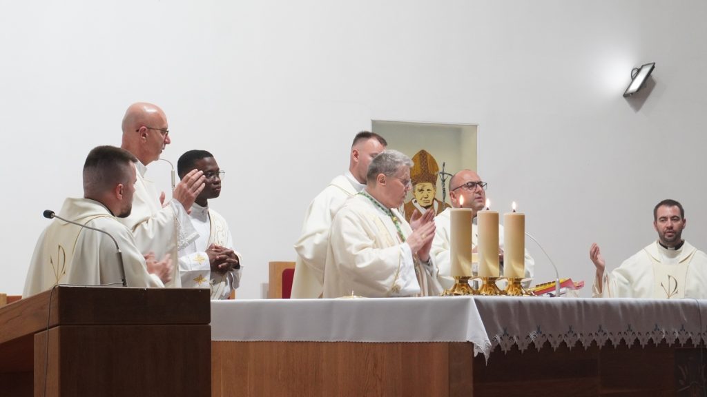 Group of priests at an altar during Mass, four tall candles lit on the cloth-covered table.