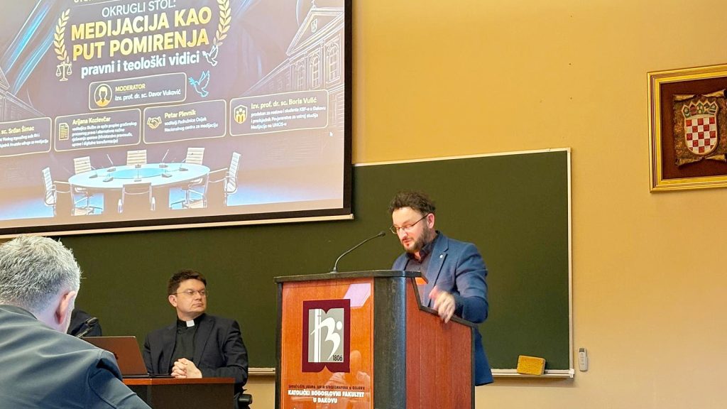 Presenter in a blue suit delivers a lecture at a wooden podium, with a panel and a large seminar slide projected behind him and a crest on the podium.