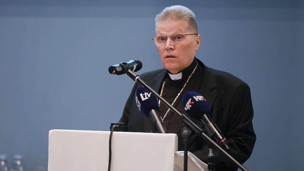 Clergyman in a clerical collar speaks at a lectern with multiple microphones during a press conference.