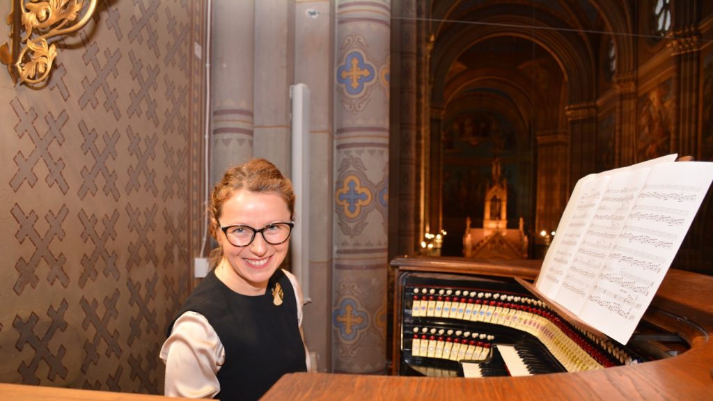 Smiling woman with glasses sits at a church organ, with sheet music on the stand in front of her.