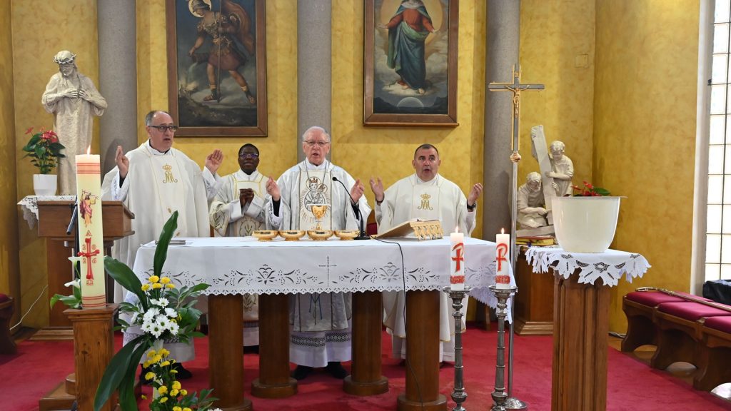 Four priests in white vestments lead a Catholic Mass at an ornate altar with lace cloth and candles inside a yellow-walled chapel.