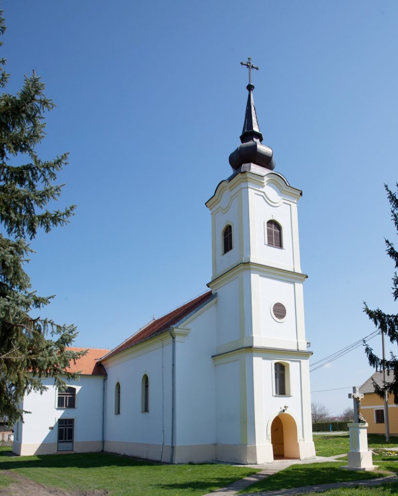 White church with a tall steeple and cross, arched windows, and an arched doorway on a green lawn against a blue sky, tree edges visible on sides.
