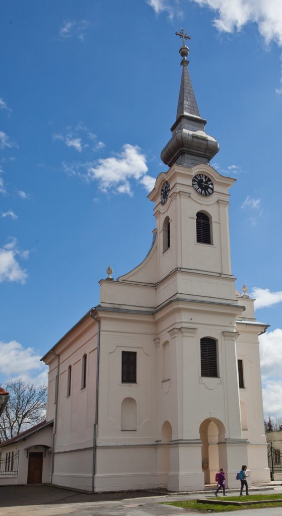 Front view of a white church with a tall steeple and clock faces, against a blue sky; two people walk toward the arched entrance
