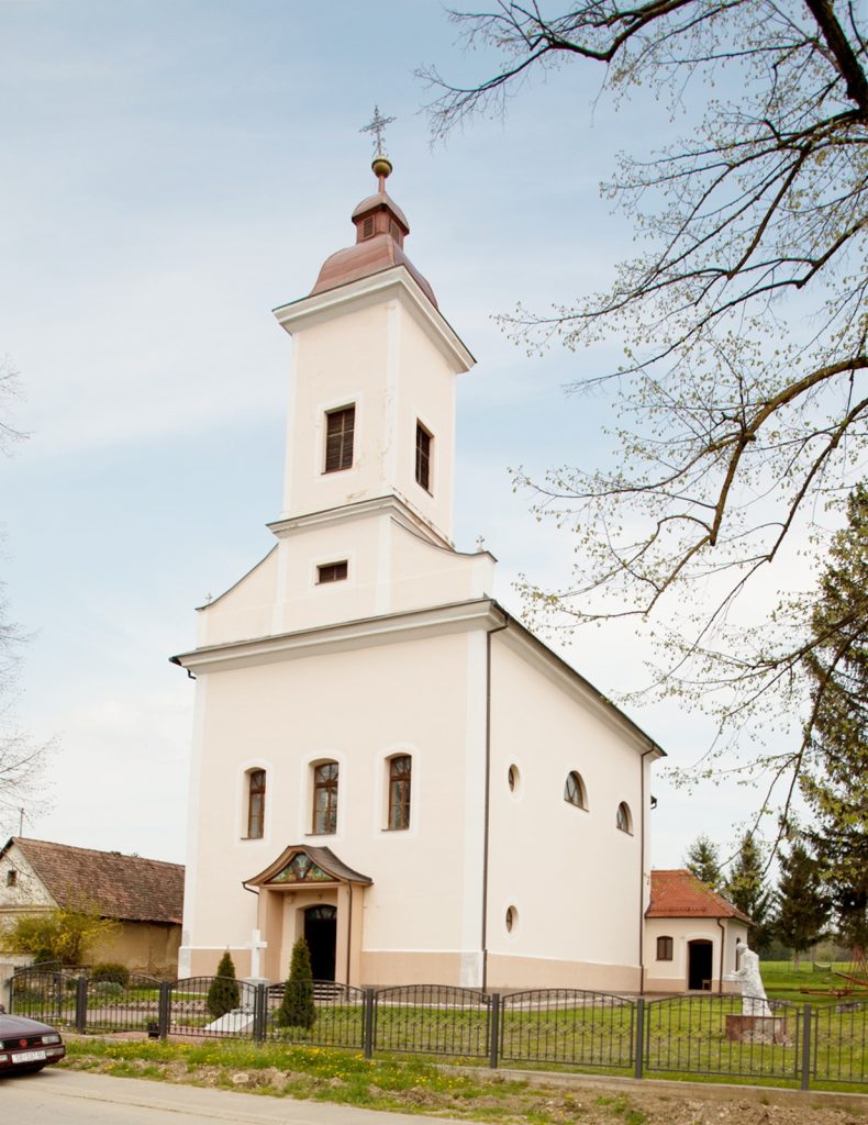 Cream-colored church with a tall square tower topped by a red roof and cross, set beside bare-branched trees and a fenced yard.