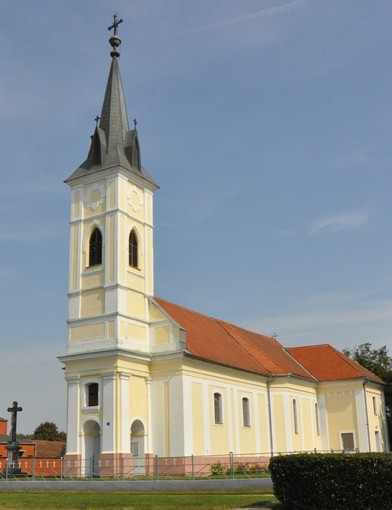 Yellow church with a tall gray steeple and cross against a blue sky; red-tiled roof visible along the side.