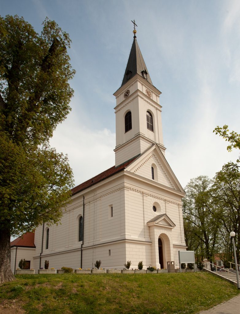 White church with a tall steeple and clock faces, surrounded by green trees on a sunny day.