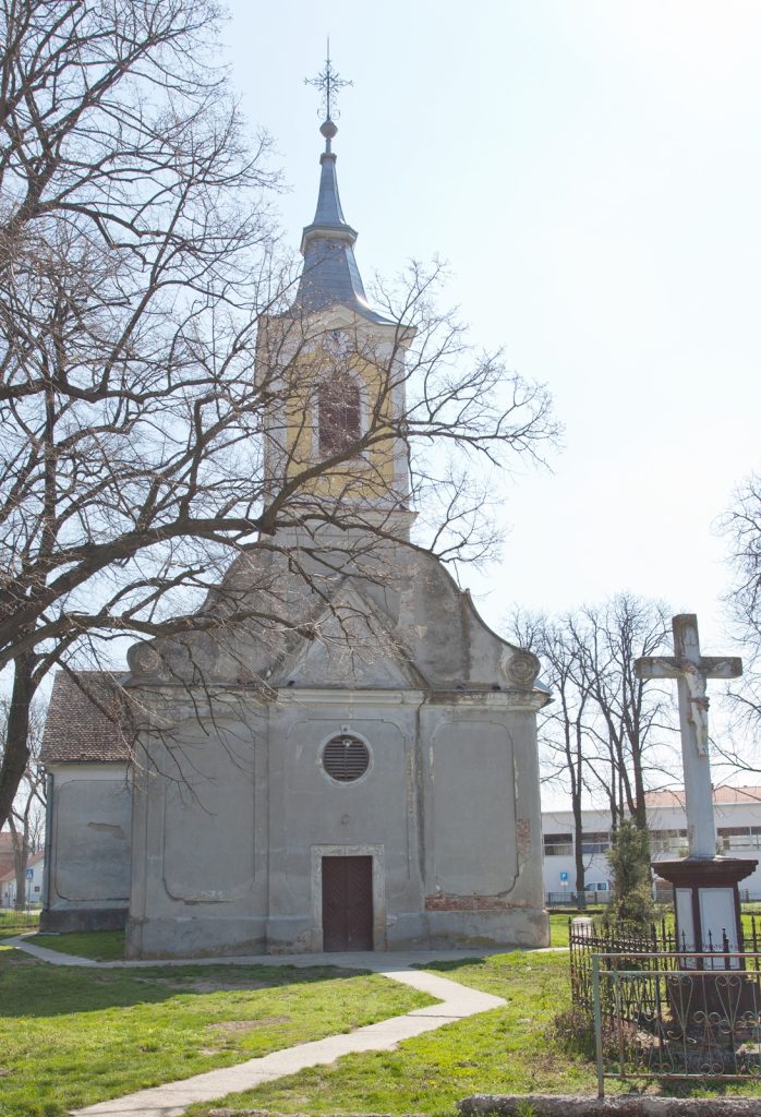 Small church with a tall steeple and weather vane, bare trees framing the facade and a curved gable roof fronting a path.