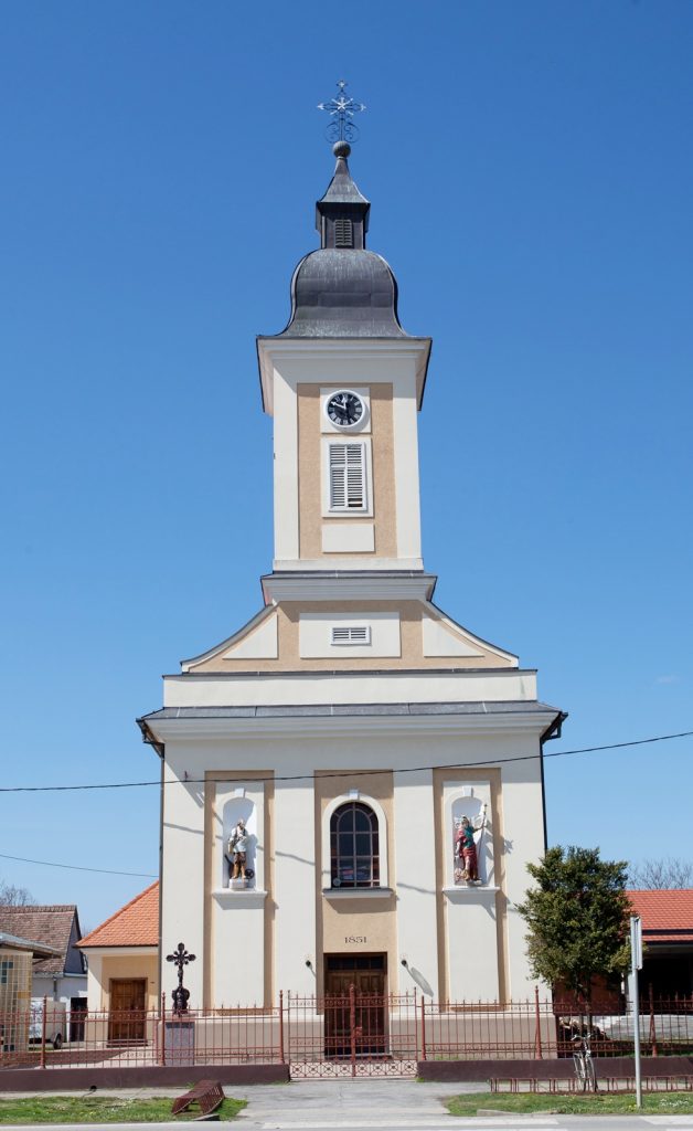 Church with tall steeple and clock tower against a blue sky; two niches with statues at the front facade.