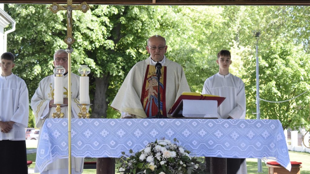 Catholic priest at an outdoor altar delivering a sermon, flanked by two altar servers, with a large cross and lit candles on the table against a green park backdrop.