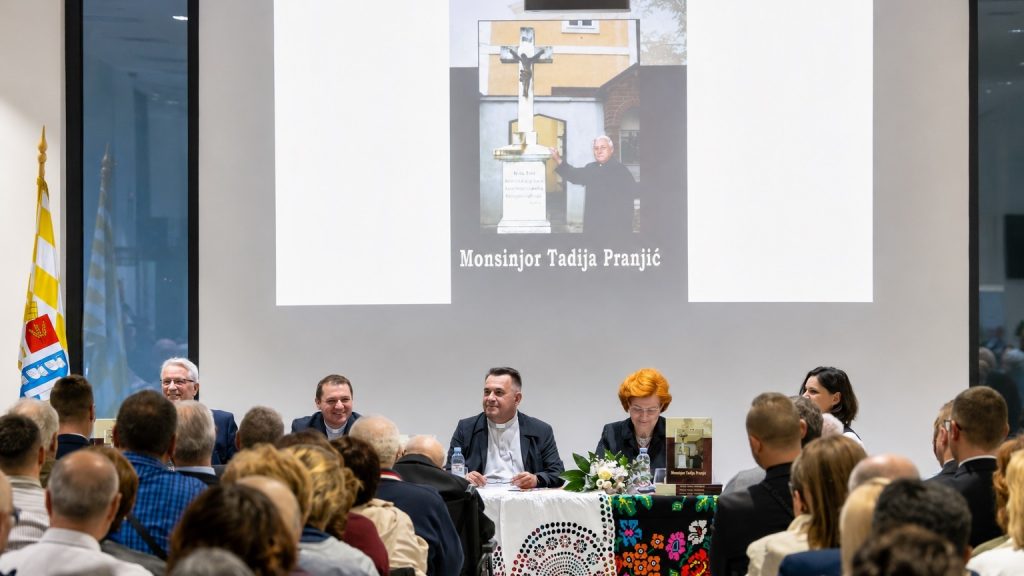 Panel of five speakers seated at a long table during a formal event, with a large projection showing Monsinjor Tadija Pranjic behind them.