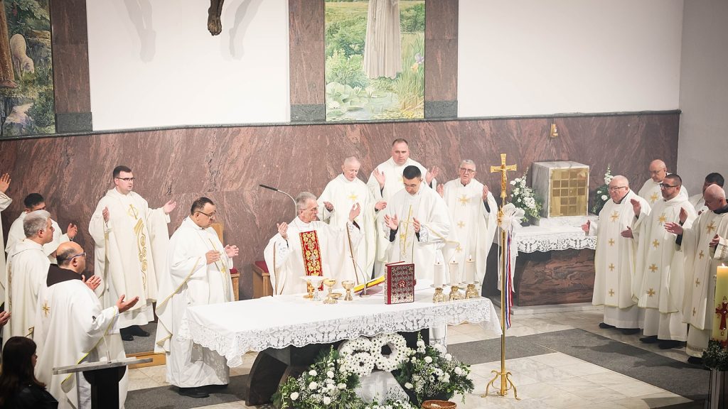 A Catholic priest and many clergy in white vestments celebrate Mass at an ornate altar with gold chalices, candles, and a rosary in front of them.