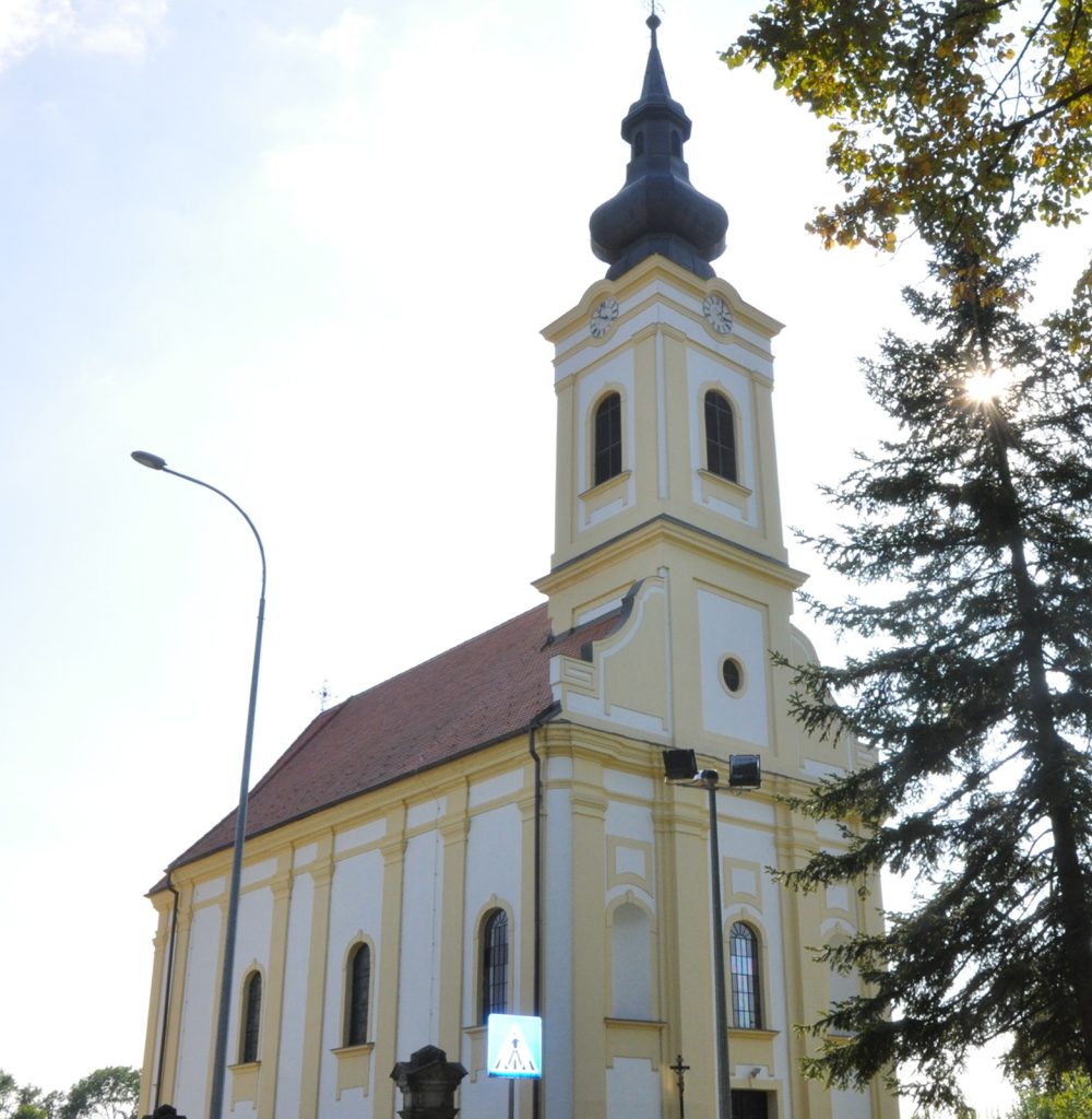 Front view of a pale yellow church with a tall onion-dome steeple, clocks on the tower, and arched windows against a bright sky.