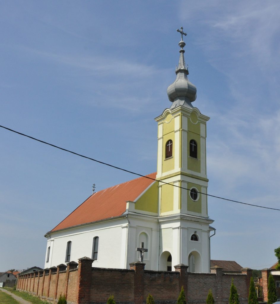 Yellow church tower with a gray onion dome and cross against a clear blue sky, red-tiled roof visible below.