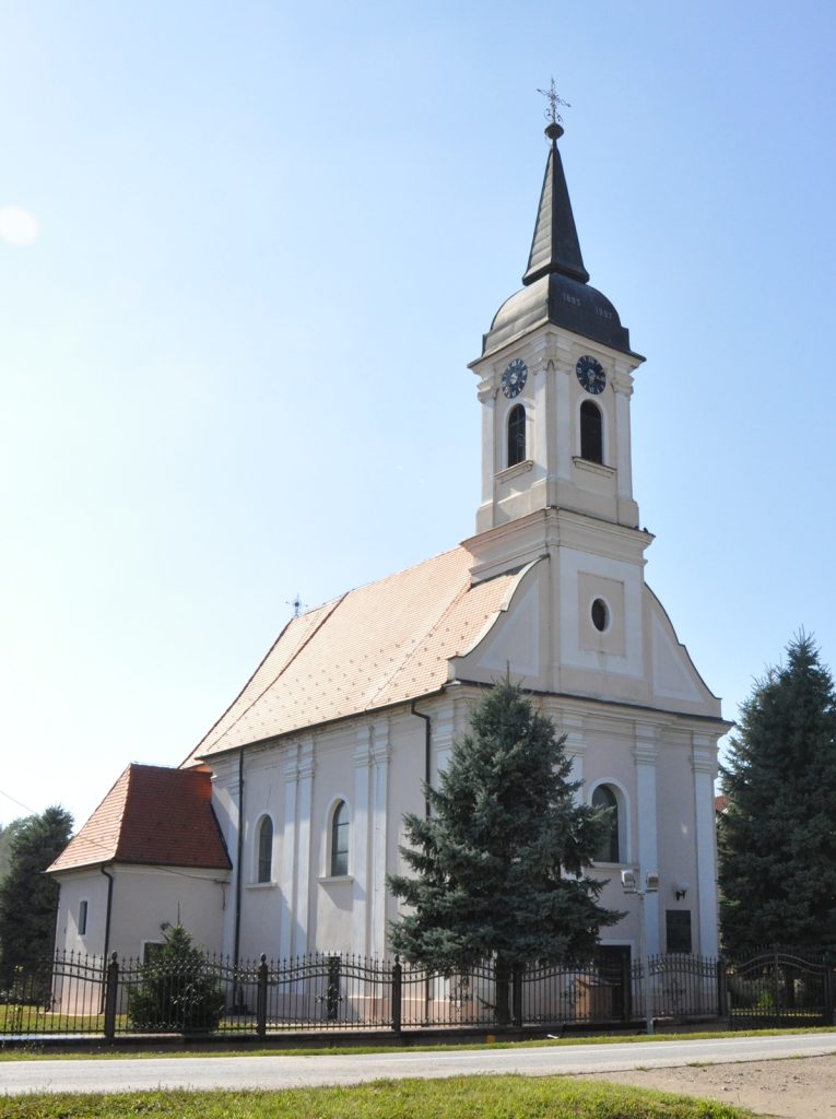 White church with a tall clock-tower and spire against a bright blue sky, surrounded by evergreens and a metal fence in the foreground.