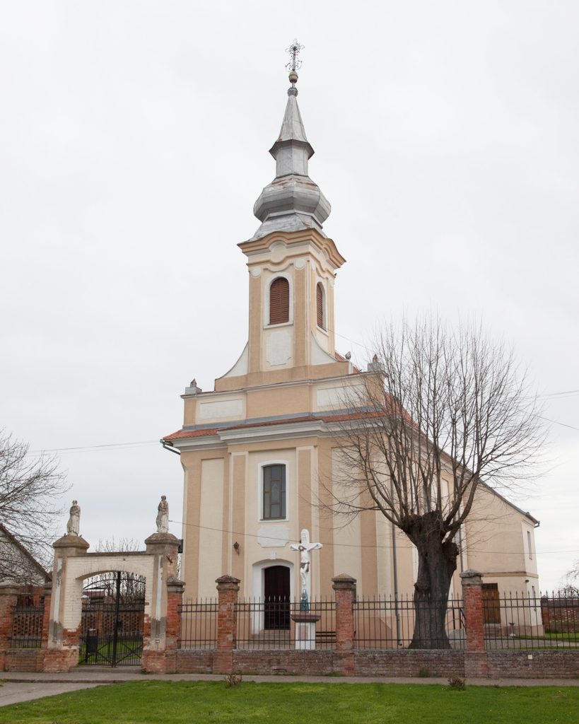 Front view of a pale-yellow church with a tall, ornate steeple topped by a cross, surrounded by a brick fence and statues at the gate on a cloudy day.