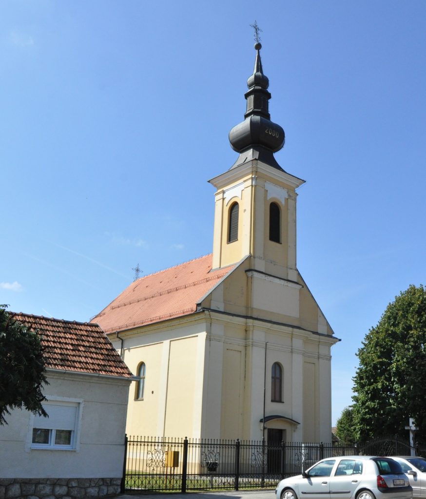 Church with pale yellow walls and a tall dark steeple rising against a clear blue sky.
