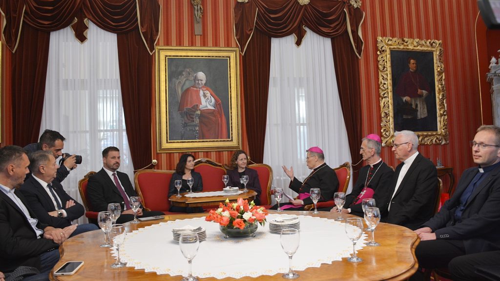 A formal meeting around a large round table in an opulent room with red drapes and two large framed portraits on the wall.