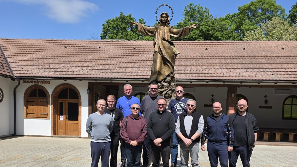 Group of men posing in front of a tall statue with a halo, outdoors in front of a building with a tile roof and wooden doors (suvenirnica sign visible).